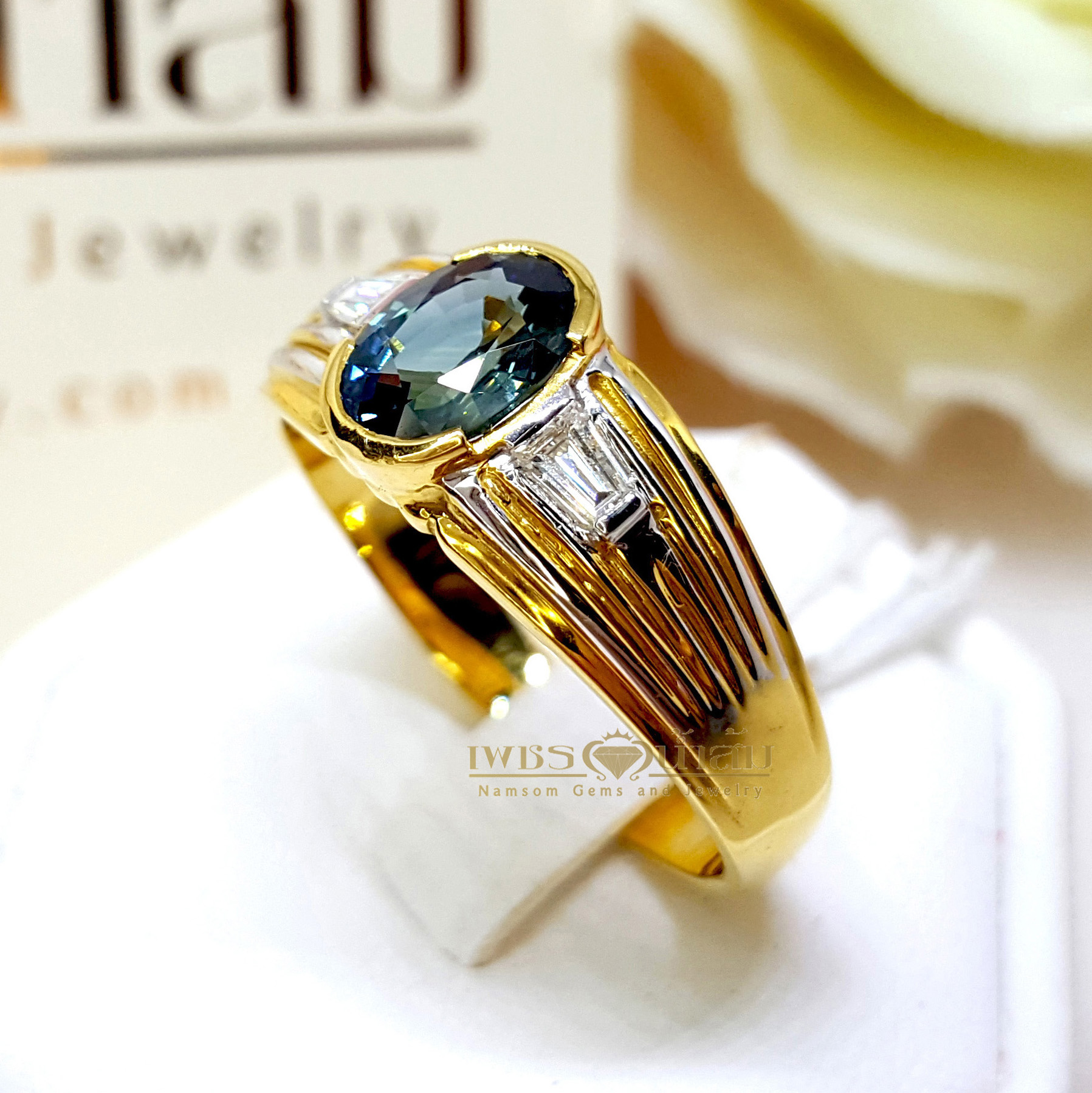 แหวนพลอยไพลิน Blue Sapphire สีอมเขียว ประดับตกแต่งด้วยเพชรแท้เจียรเหลี่ยมแบเก็ต และบากลายทางที่ขอบข้าง