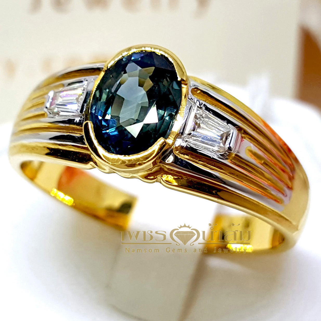 แหวนพลอยไพลิน Blue Sapphire สีอมเขียว ประดับตกแต่งด้วยเพชรแท้เจียรเหลี่ยมแบเก็ต และบากลายทางที่ขอบข้าง